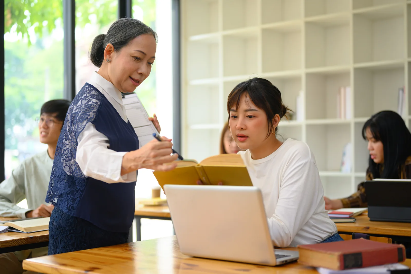 A tutor helping a student work on a laptop in a classroom