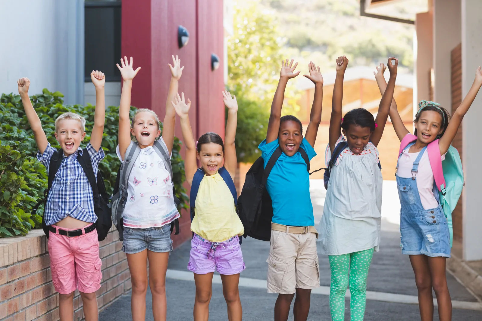 Elementary students standing in a row on a school campus with their hands raised